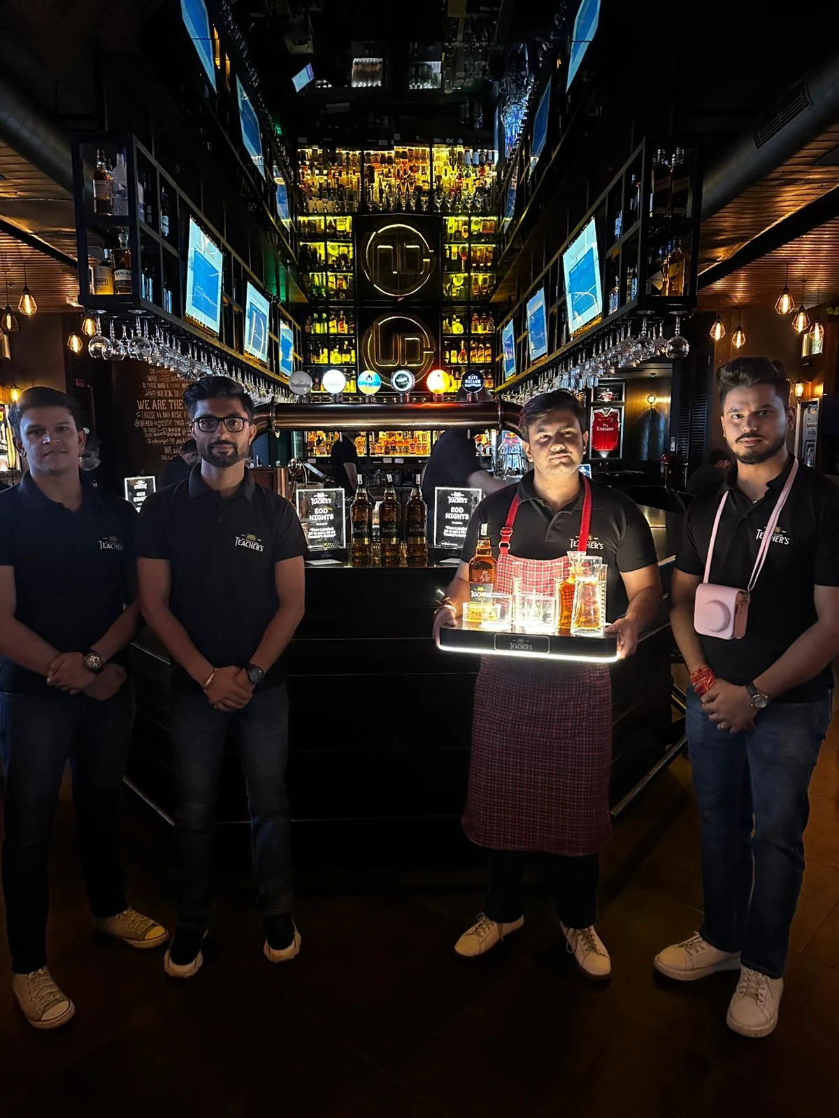 Teacher's bar team in branded polos at the glowing back bar, bartender with illuminated serve tray